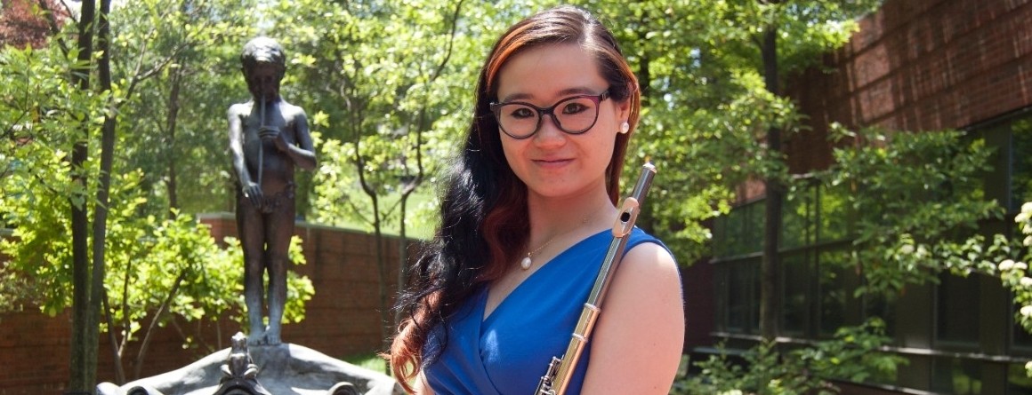 A graduate student poses with her flute in front of a fountain on campus.