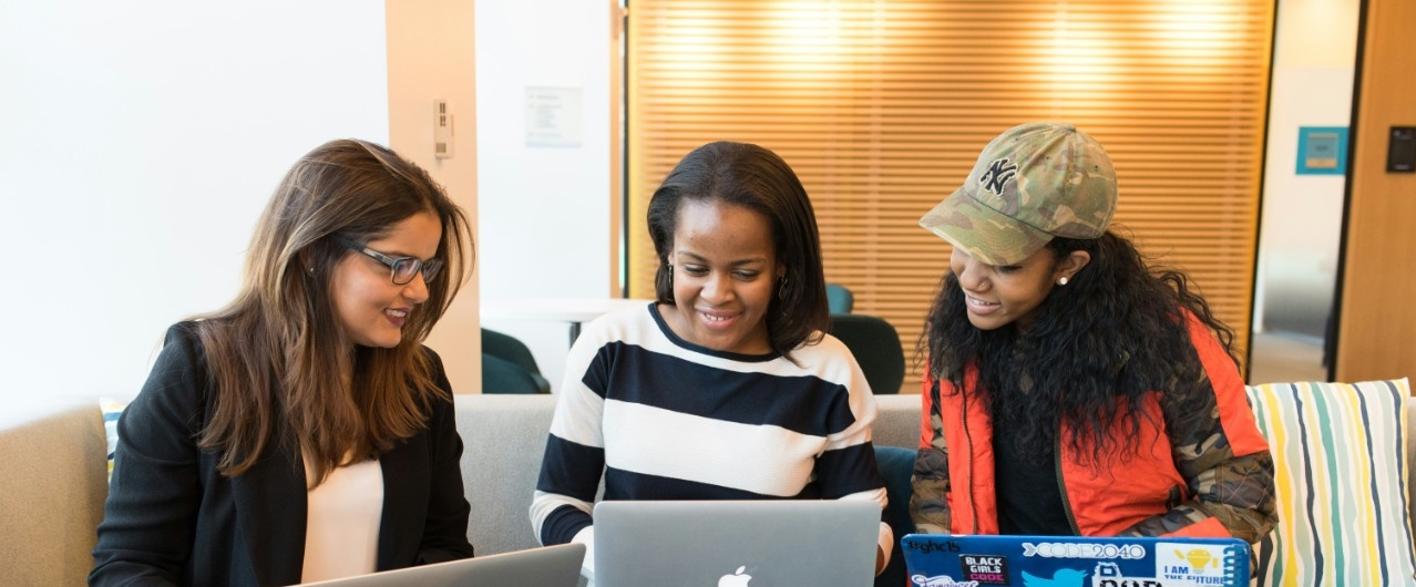 Three students sit together on a couch with their laptops and talk.